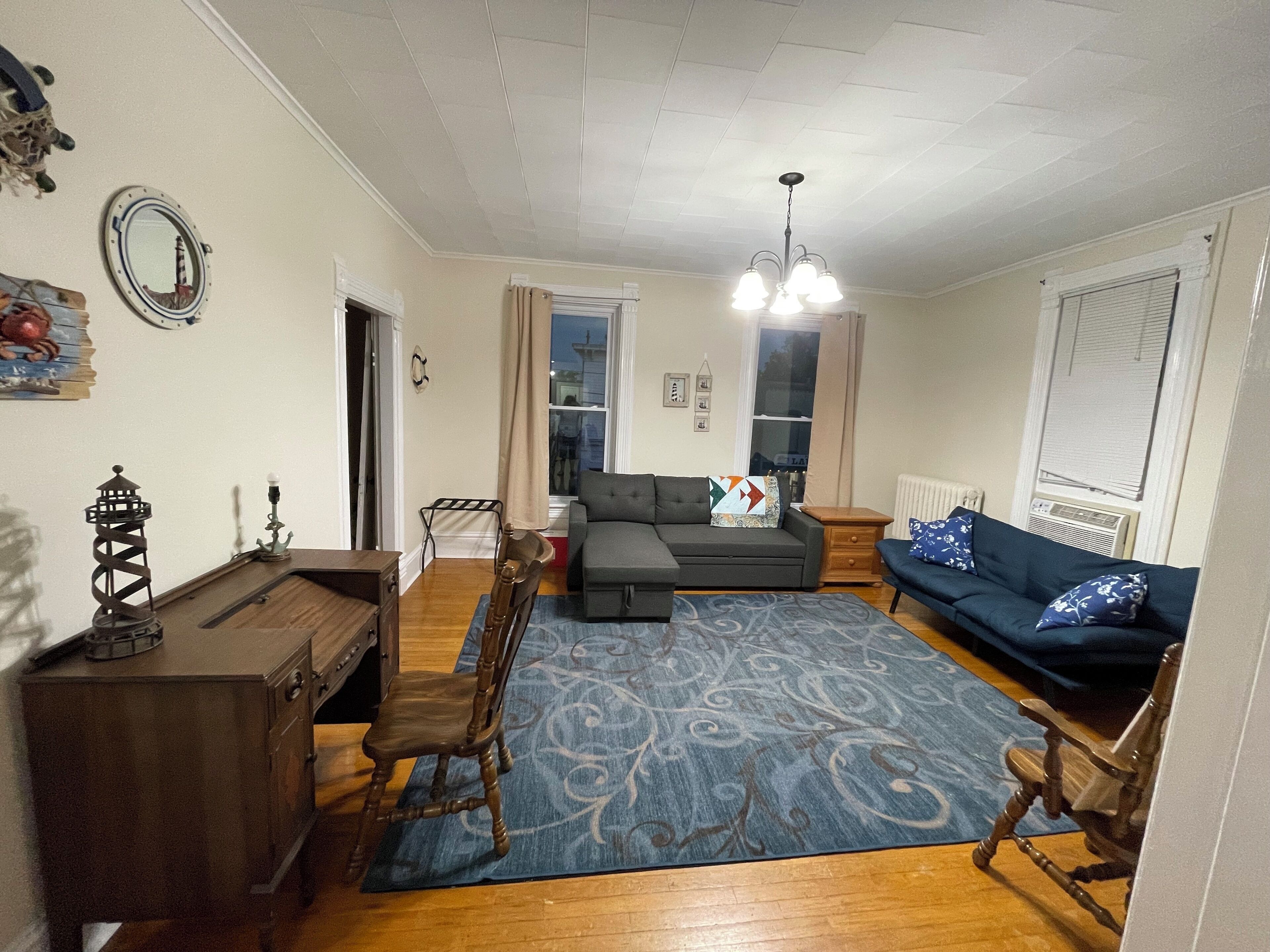 A living room with a sofa, a dining table with chairs, a chandelier, and a patterned rug.