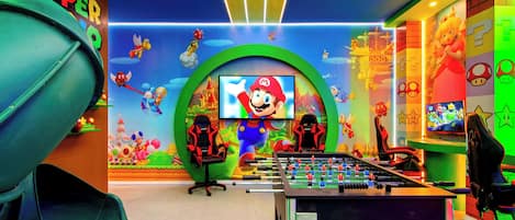Una habitación luminosa con una mesa de fútbol de mesa, dos sillas de juego y una pantalla grande mostrando un personaje de videojuego.