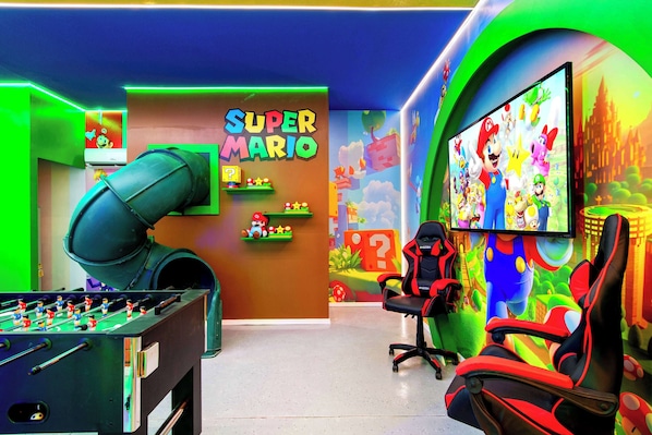 A gaming room with a foosball table, a large screen, and a Super Mario themed wall.