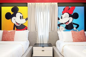 A hotel room with two beds, a nightstand, and Mickey Mouse and Minnie Mouse paintings on the walls.