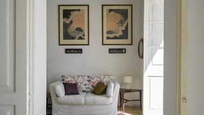 Ein Wohnzimmer mit einem weißen Sofa, floralen Kissen, zwei gerahmten Kunstwerken an der Wand und einem kleinen Tisch mit einer Lampe.