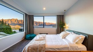 Un dormitorio con un ventanal grande que ofrece vistas a la montaña, una cama con ropa blanca, un sillón informal de color verde y una mesita de noche con una lámpara.