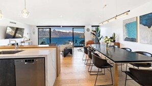 A modern kitchen with a dishwasher, a dining area with a long table and chairs, and a large window offering a view of mountains and a lake.
