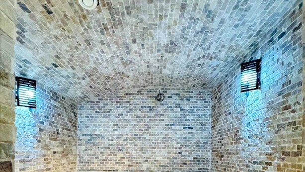 Habitación con pared de ladrillo y piso de baldosas, que incluye un banco y una rejilla de ventilación en el techo.