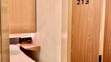 A compact hotel room with a bed, a small bedside table, a built-in shelf, and a wooden wardrobe with the number 213.