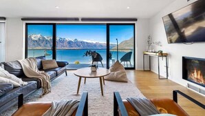 A modern living room with a large window offering a mountain and lake view, a fireplace, and a flat-screen TV mounted on the wall.