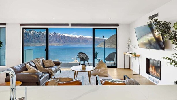 A modern living room with a large window offering a view of a lake and mountains. There is a leather sofa, a small round table, and a flat-screen TV mounted on the wall.