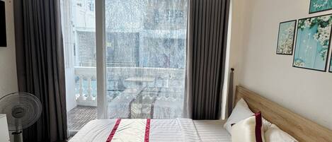 A hotel room with a large bed, a standing fan, a balcony with curtains, and a view of the cityscape.