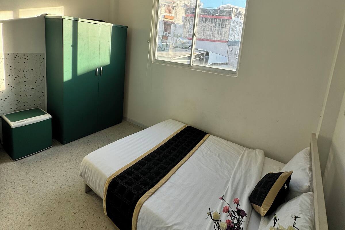 A bedroom with a bed, a green wardrobe, a small table, and a window with a view of buildings.