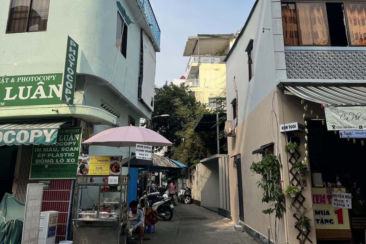 A narrow street with buildings on either side, including a shop with a sign for 'Luân Photocopy'.