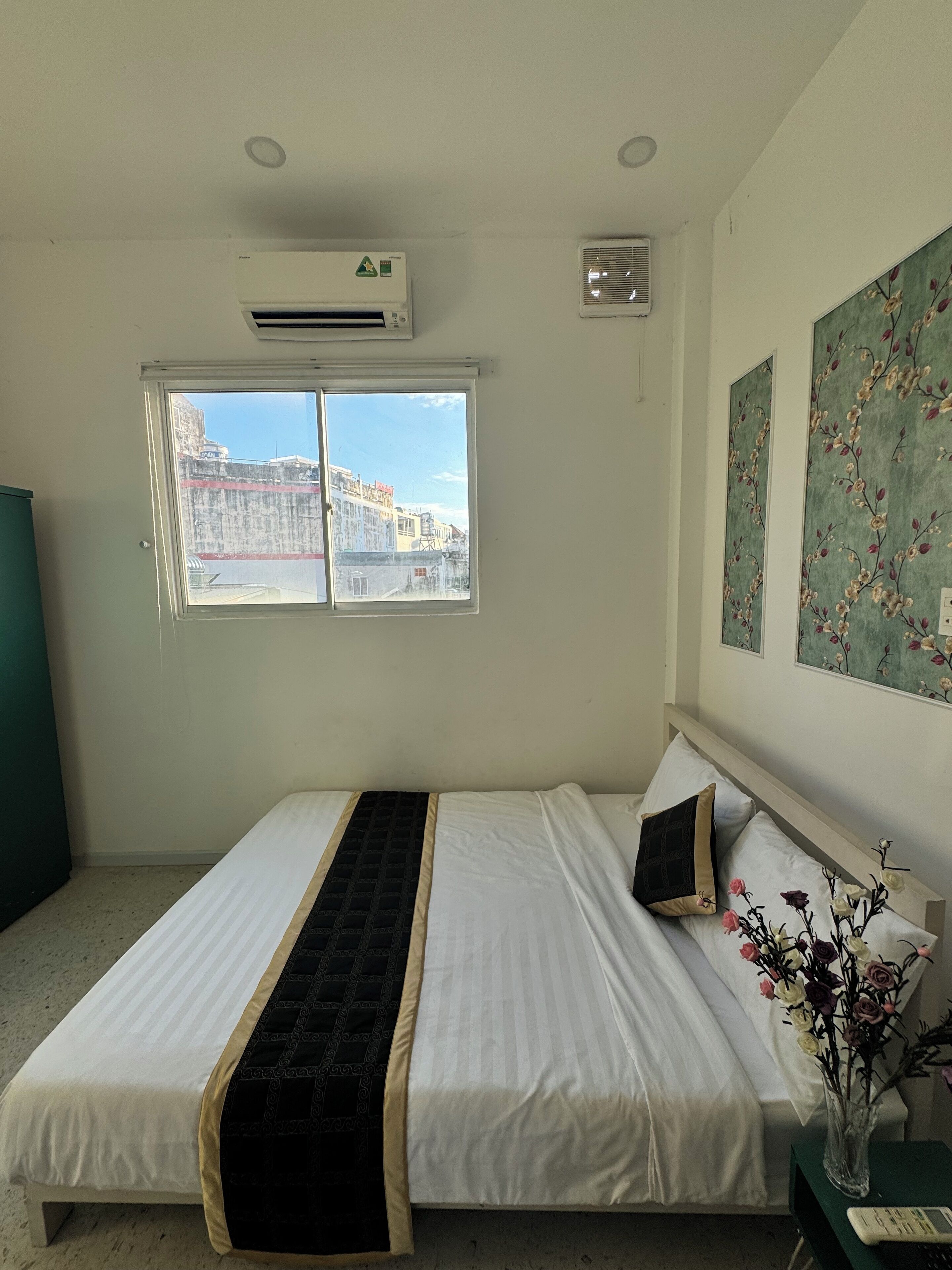 A bedroom with a bed, a window, an air conditioning unit, and a vase with flowers.