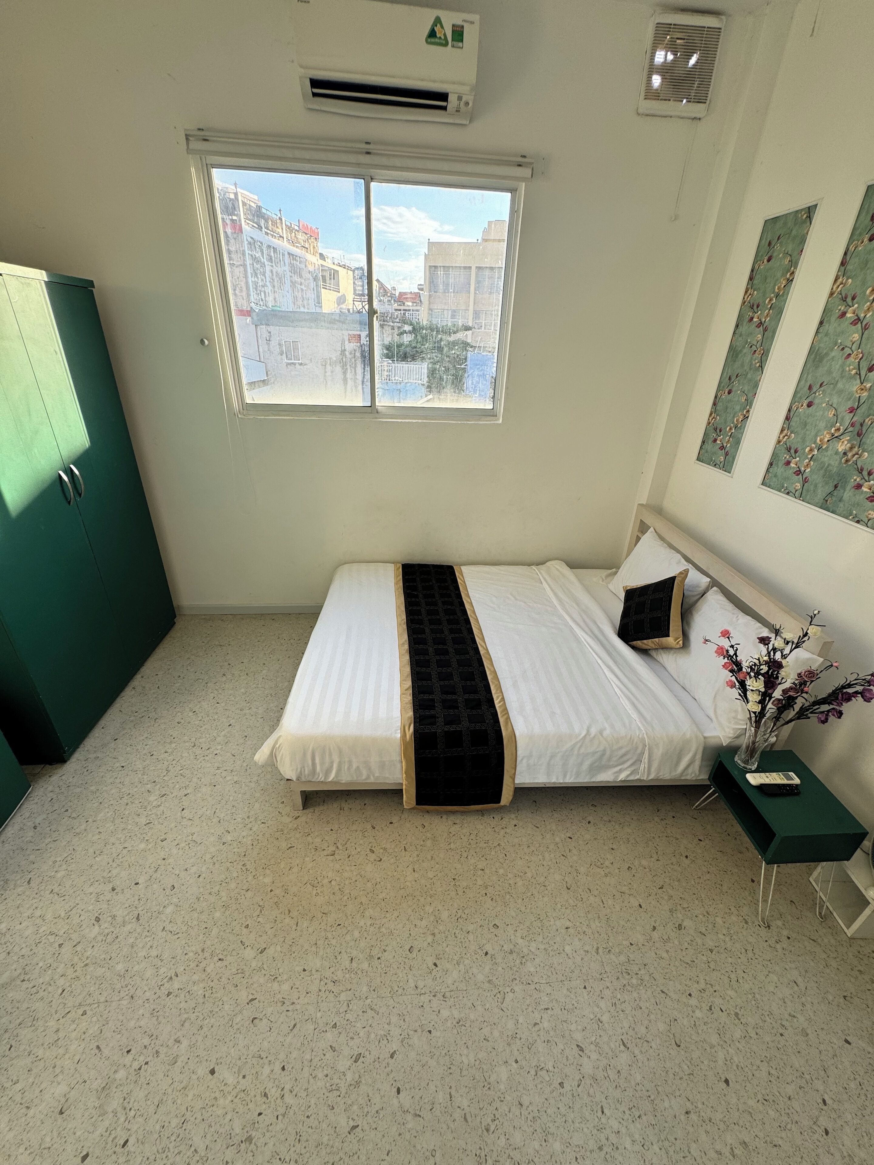 A bedroom with a bed, a window, a green cabinet, a small table with a vase of flowers, and a wall-mounted air conditioner.