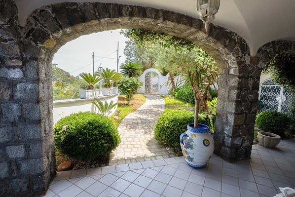 An arched stone entrance with a tiled pathway leading to a white gate, surrounded by greenery and potted plants.
