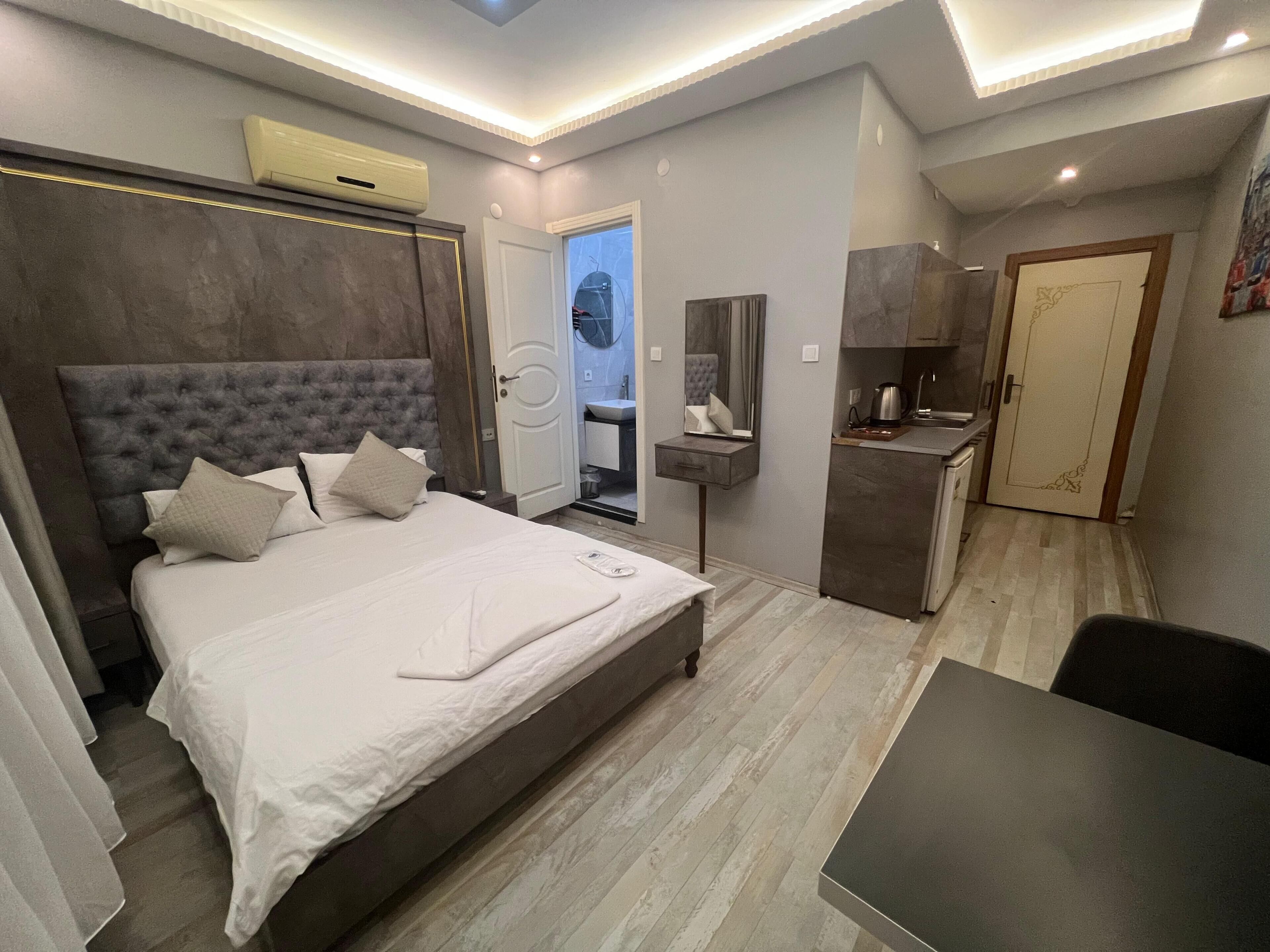 Ein modernes Hotelzimmer mit einem großen Bett, einem Schreibtisch, einem Spiegel, einer Küchenzeile und einem sichtbaren Badezimmer durch eine geöffnete Tür.