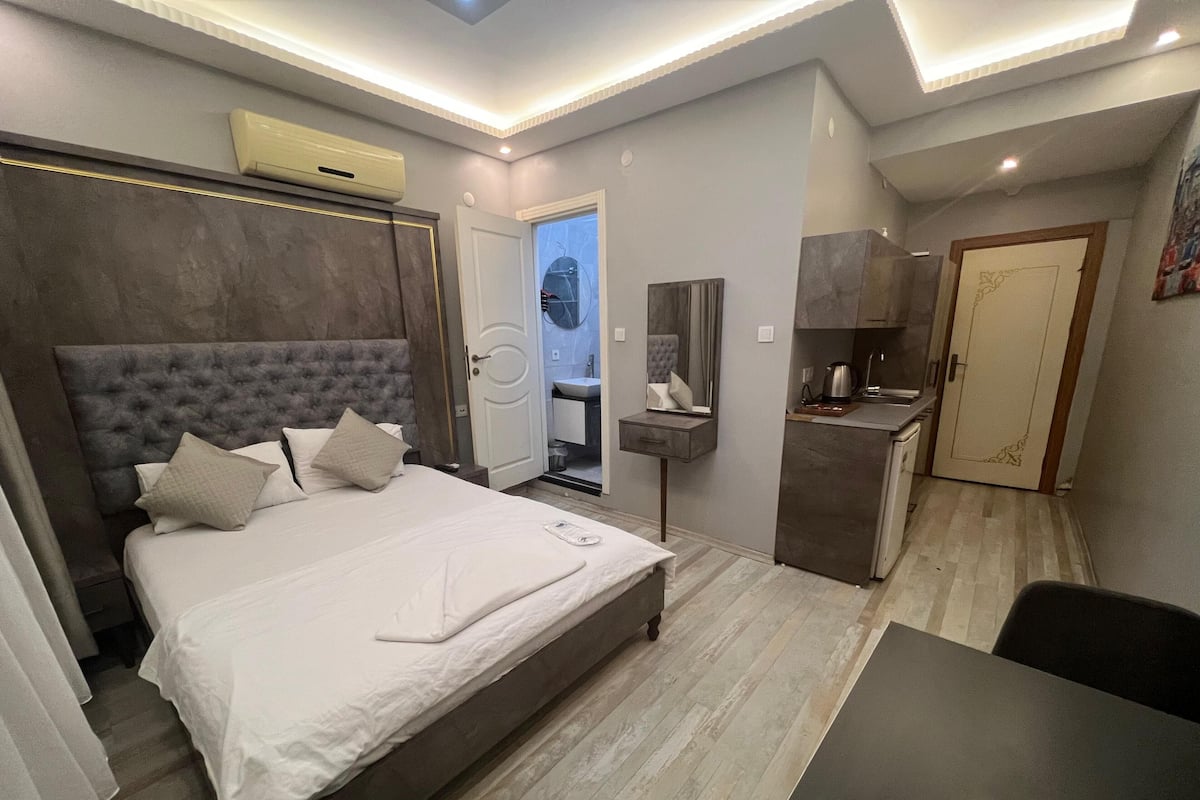 A modern hotel room with a large bed, a desk, a mirror, a kitchenette, and a bathroom visible through an open door.
