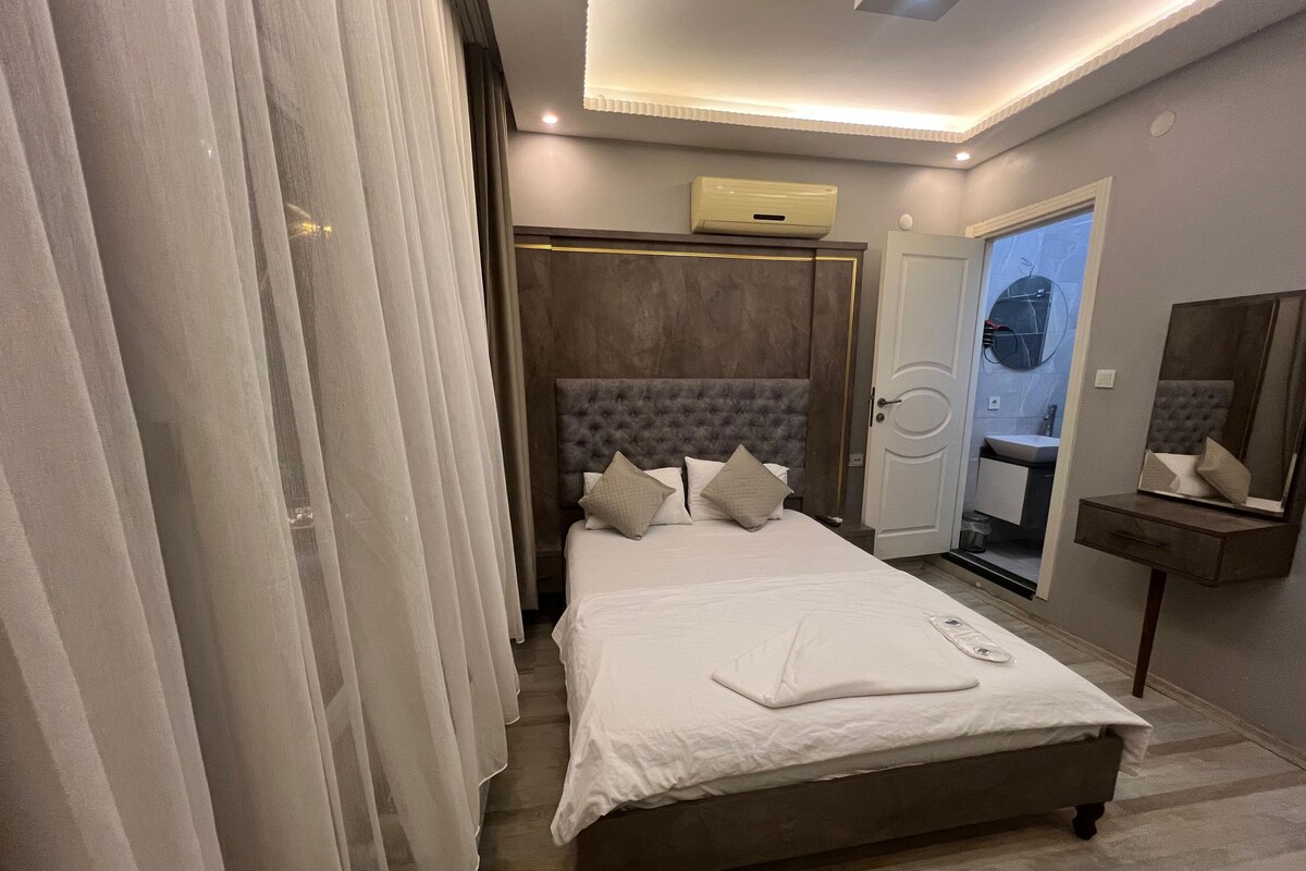 A modern bedroom with a large bed, a bedside table, a mirror, and a bathroom visible through an open door.