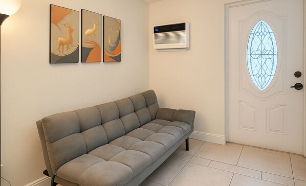 Ein Wohnzimmer mit grauem Sofa, zwei an der Wand befestigten abstrakten Gemälden, einer Wand-Klimaanlage und einer Tür mit geometrischem Glasdekor.