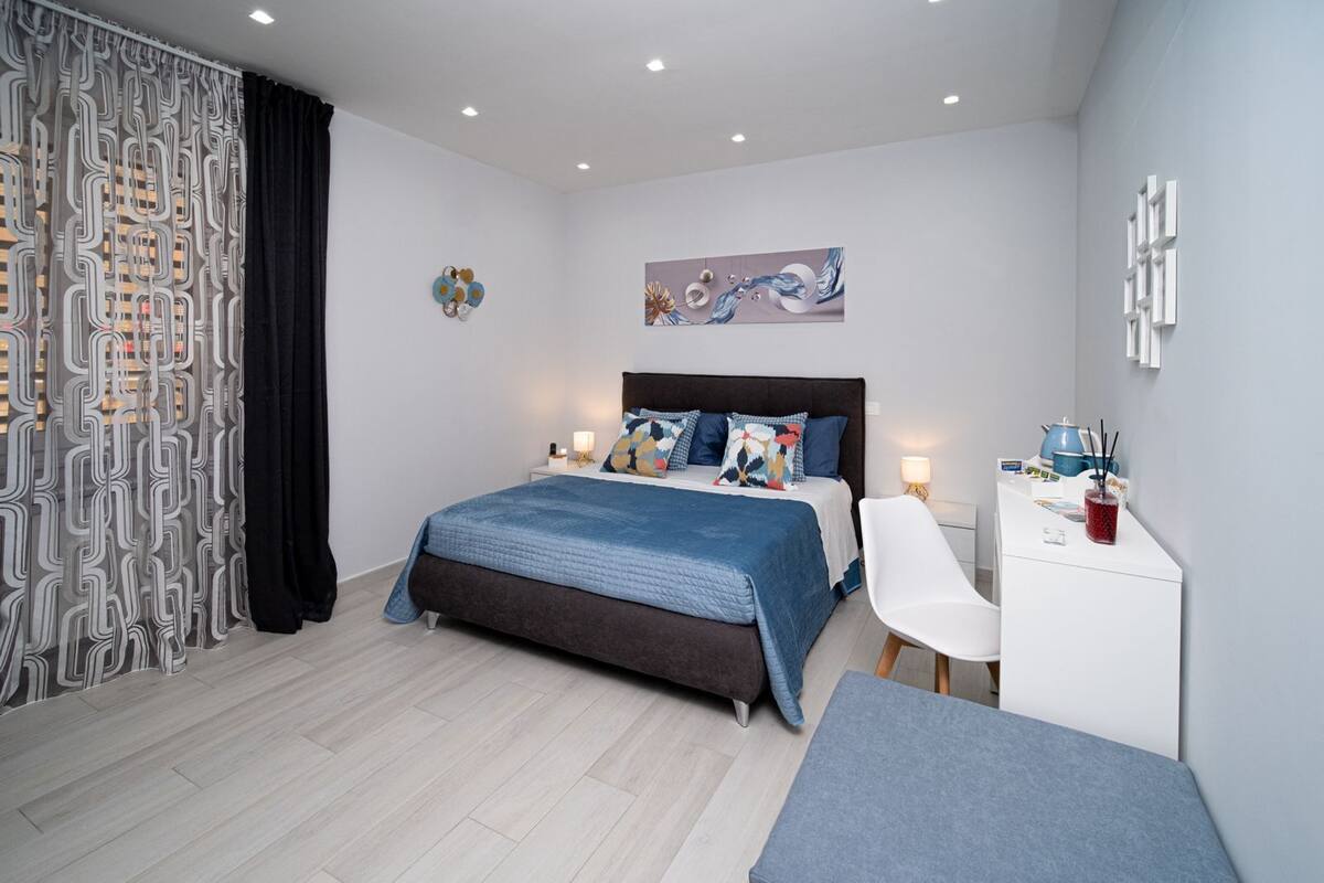 Une chambre à coucher moderne, comprenant un lit, une table de chevet, une chaise, un bureau et une œuvre d’art.