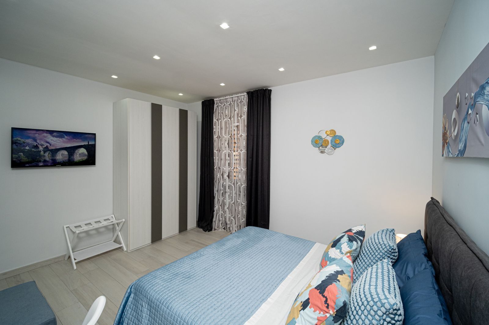 A modern bedroom with a bed, a small white table, a wall-mounted TV, and a decorative wall piece.