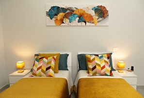 Habitación de hotel con dos camas, cada una con una colcha amarilla, un cojín estampado y una mesita de noche con lámpara. Una obra de arte colorida adorna la pared sobre las camas.