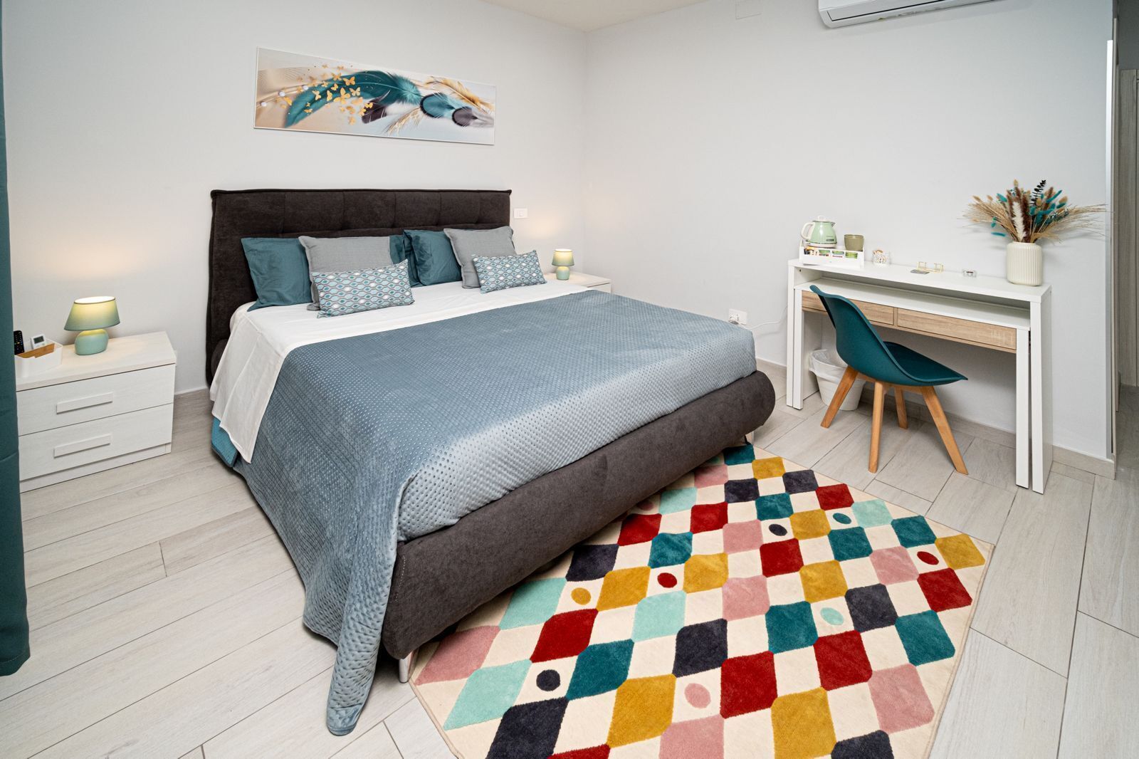 A bedroom with a bed, bedside table, desk, chair, and a colorful rug.