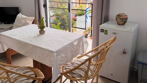 A room with a table, two wicker chairs, a TV, a small refrigerator, and a view of a balcony with plants and buildings. - Monoambiente El Arrayán (Echeverry)