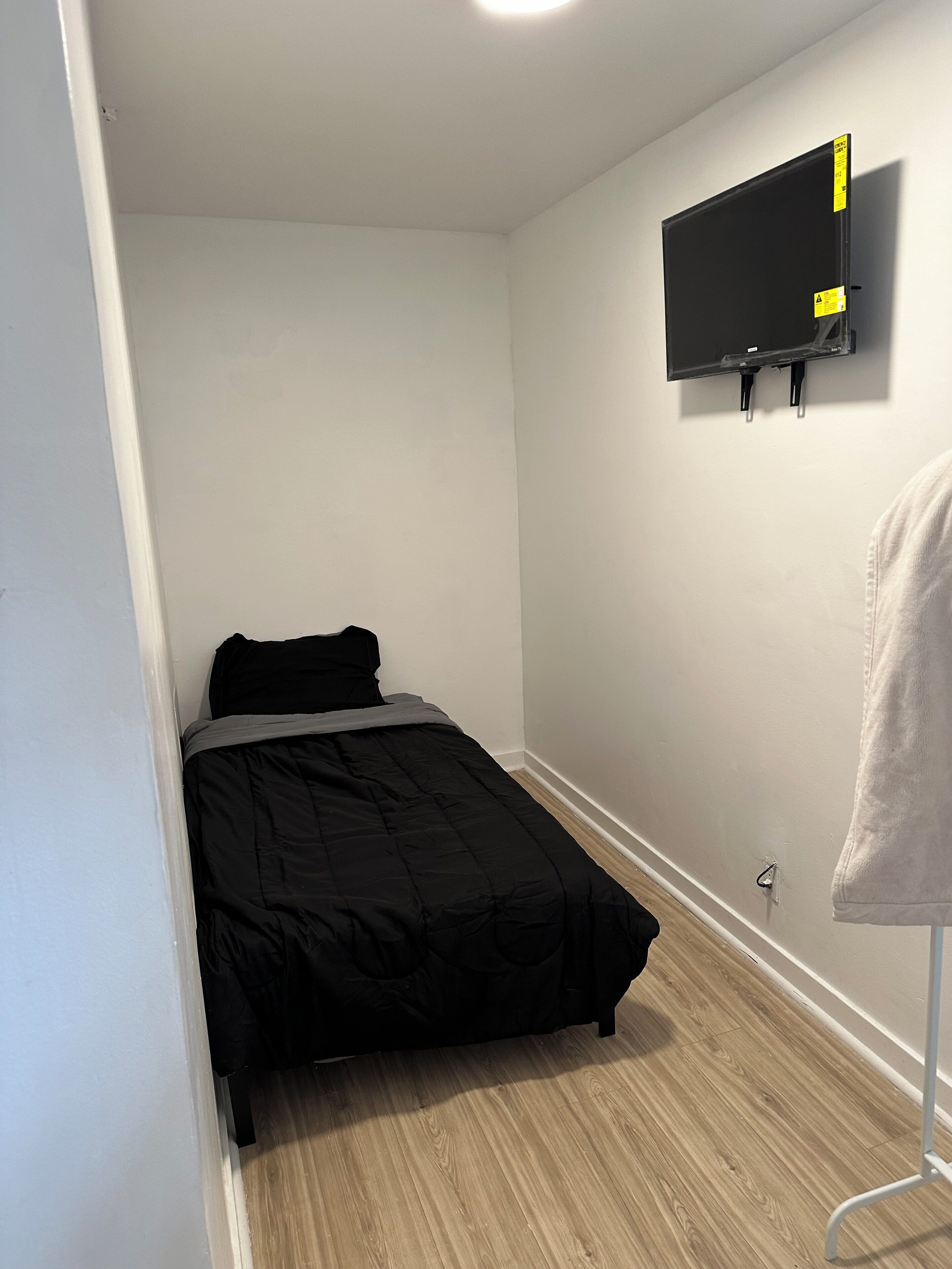 Una habitación pequeña y poco amueblada con una cama, un televisor montado y un perchero.