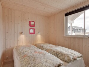 A room with wooden walls, a bed with patterned bedding, two wall-mounted lights, and a window with a view of a building outside.