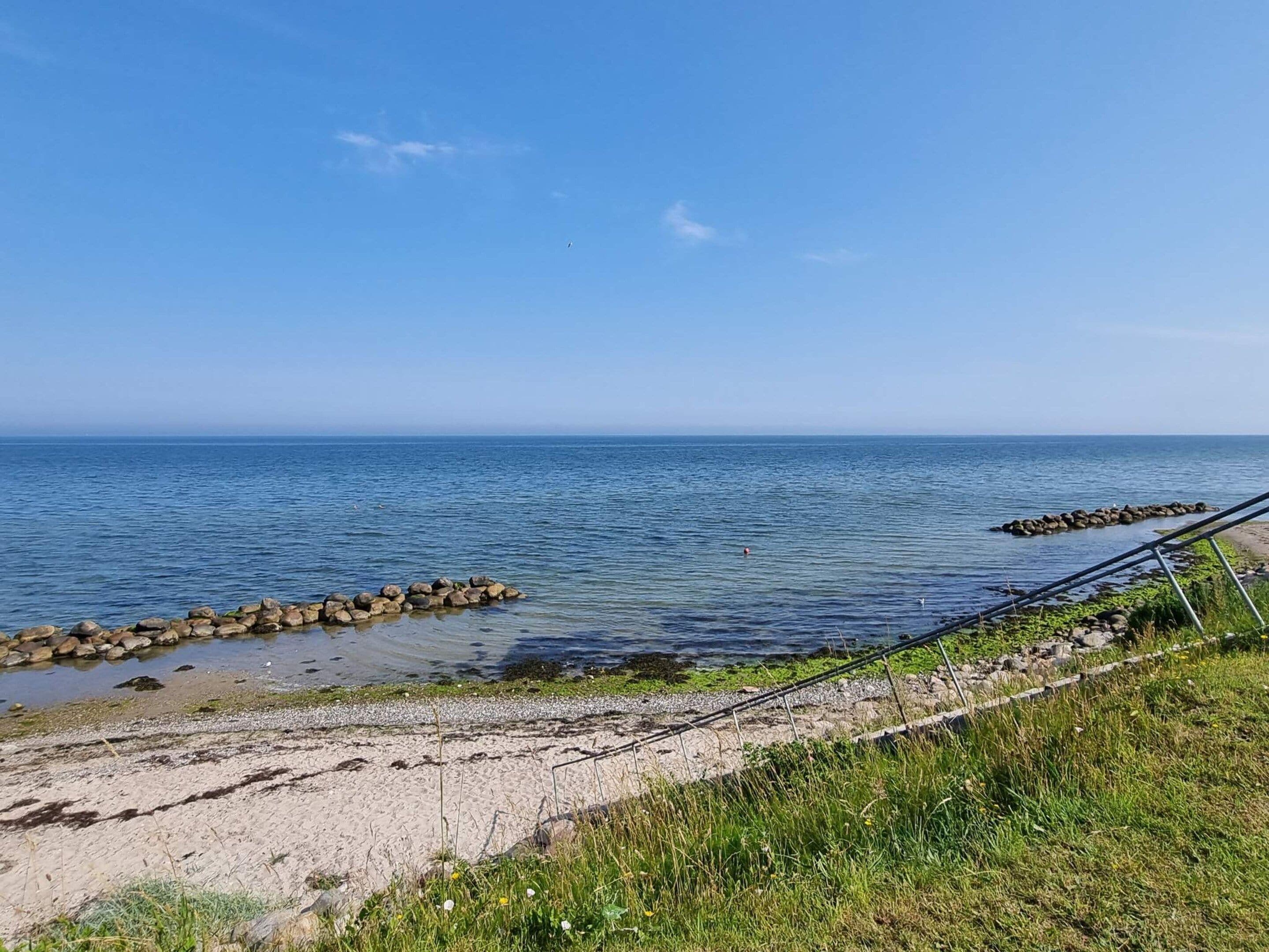 A coastal landscape with a sandy beach, a rocky breakwater, and a clear blue sky.
