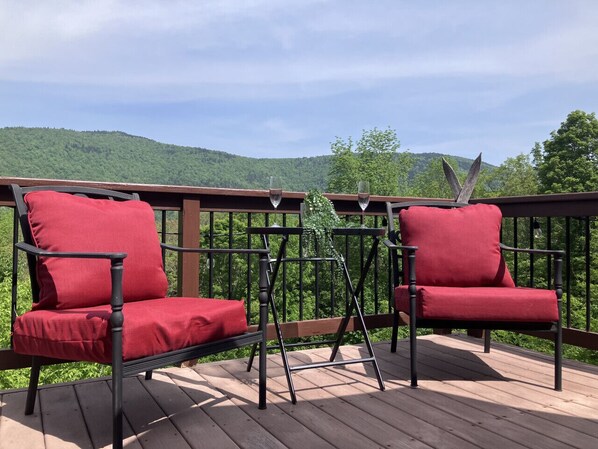 A wooden deck with two red cushioned chairs and a small table, overlooking a forested hillside.