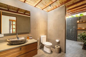 A modern bathroom with a stone sink, wooden vanity, a toilet, and a shower area. - Villa Nya Ubud (Ubud)