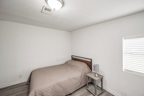 Ein Schlafzimmer mit Bett, Nachttisch, Lampe, Fenster mit Jalousien und Deckenleuchte.