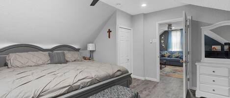 Un dormitorio con cama, una banca, ventilador de techo, cómoda y una puerta que da a otra habitación.