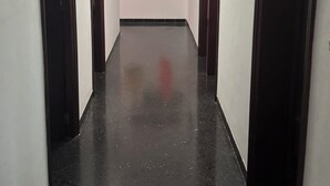 A hallway with a polished black floor, white walls, and a ceiling light. There are doors on both sides, a wall-mounted air conditioner, and a shelf with hats and bags.