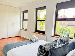 A bedroom with a bed, pillows, a radiator, and windows with a view of greenery and buildings.
