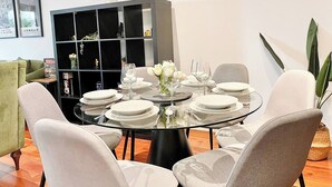 Spiseområde med et rundt glasbord dækket til seks personer, omgivet af fire stole. En sort reol med pyngegenstande står op ad væggen.