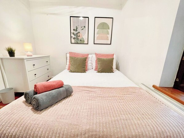 A bedroom with a bed, pillows, a bedside table with a lamp, a small plant, and artwork on the wall.