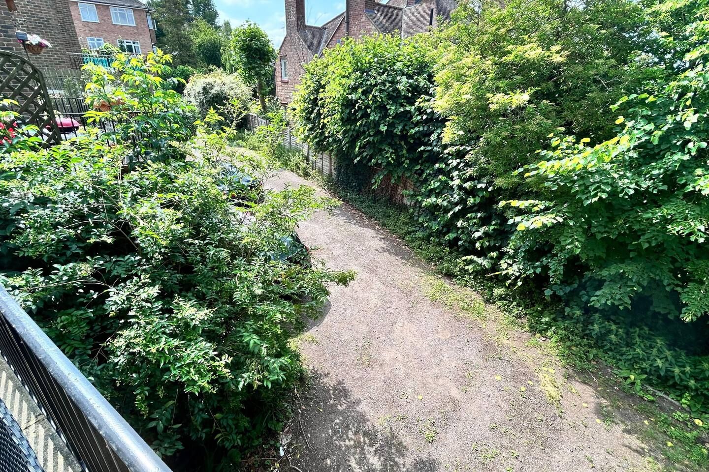 A narrow, unpaved path flanked by dense greenery and a brick building in the background.