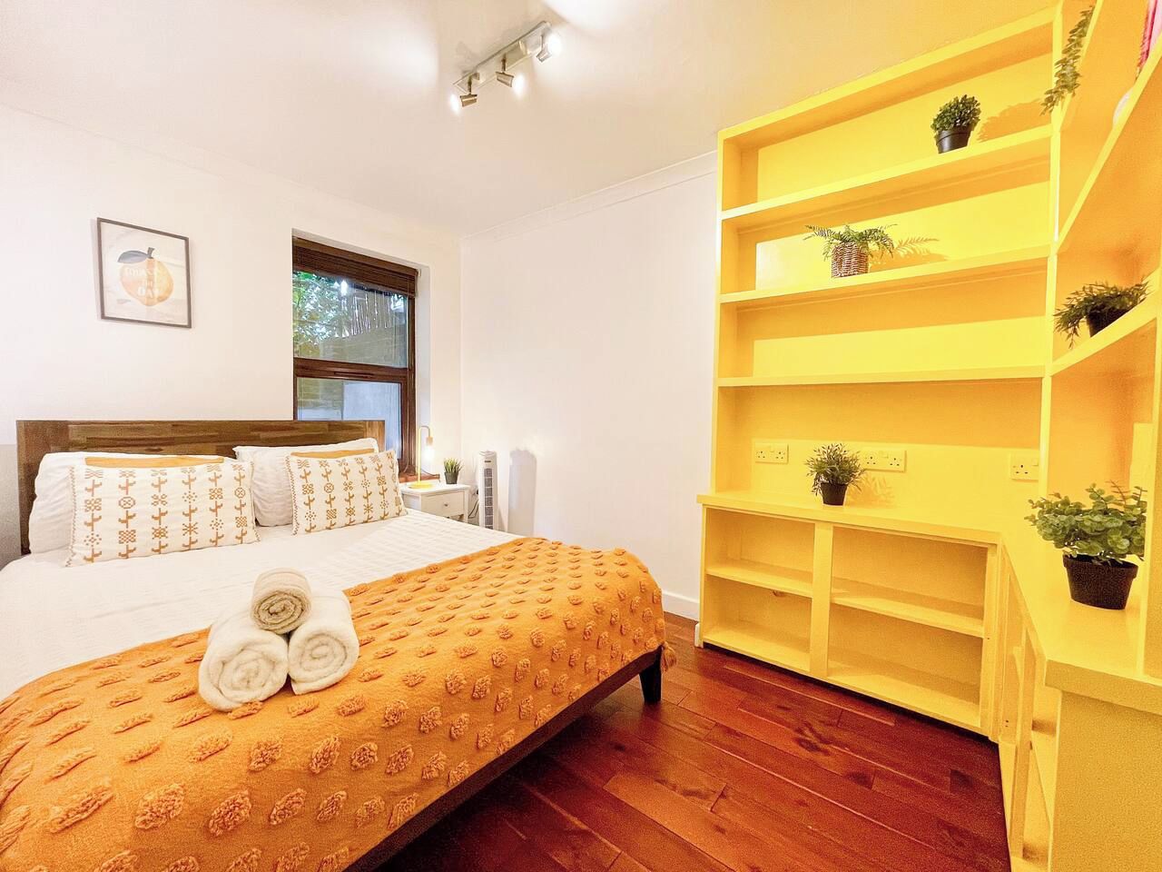 Une chambre d’hôtel avec un lit, une tête de lit en bois, une fenêtre, une étagère jaune avec des plantes et un tableau accroché au mur.