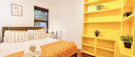 Une chambre d’hôtel avec un lit, une tête de lit en bois, une fenêtre, une étagère jaune avec des plantes et un tableau accroché au mur.