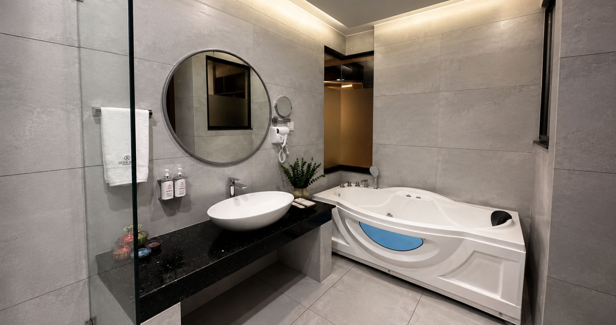 Un baño moderno con un espejo redondo, un lavamanos blanco, una bañera y una planta pequeña en la encimera.