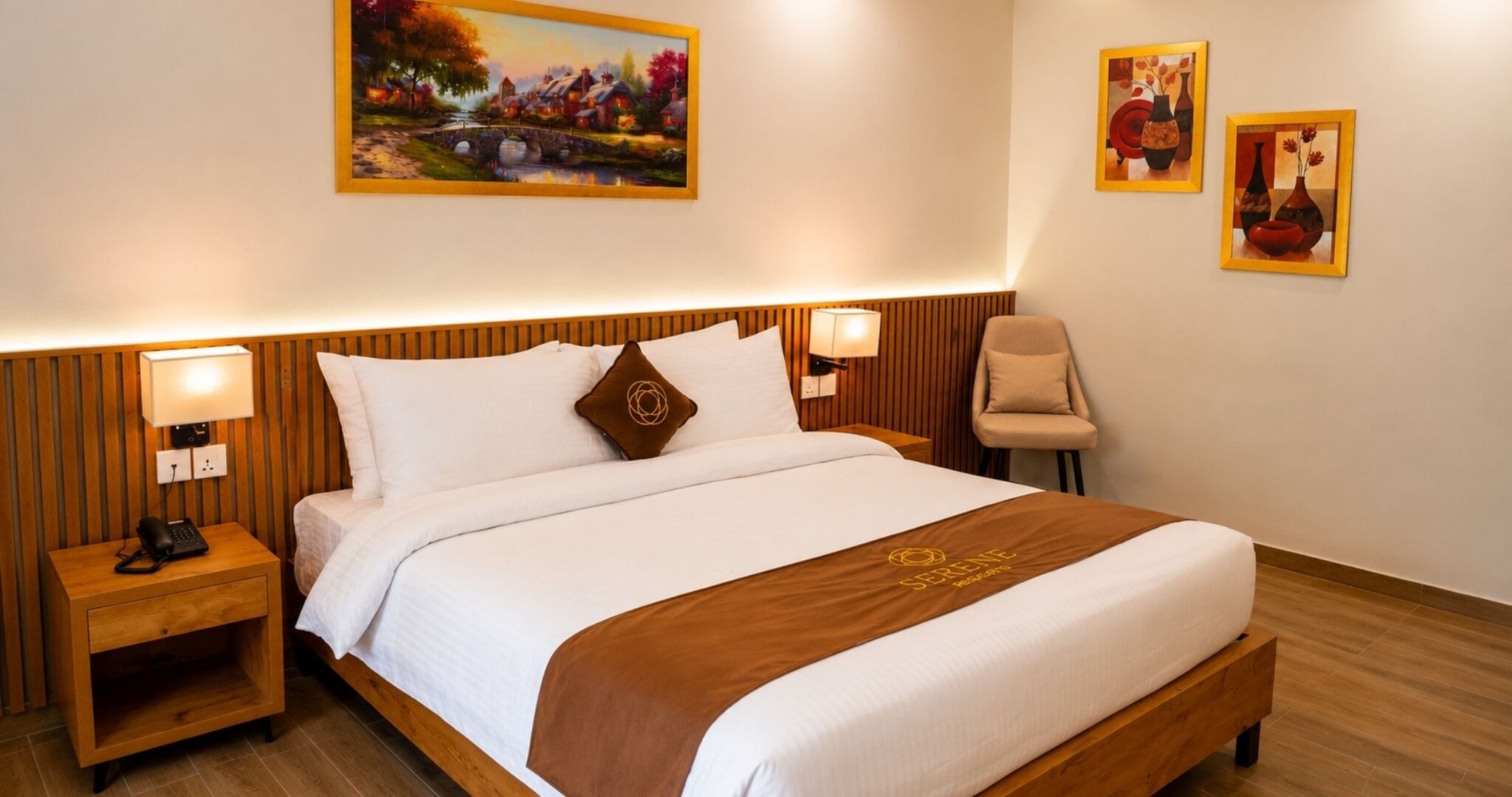 A hotel room with a wooden headboard, a bed with white bedding, a brown throw, two bedside lamps, a nightstand with a phone, and two framed paintings on the wall.