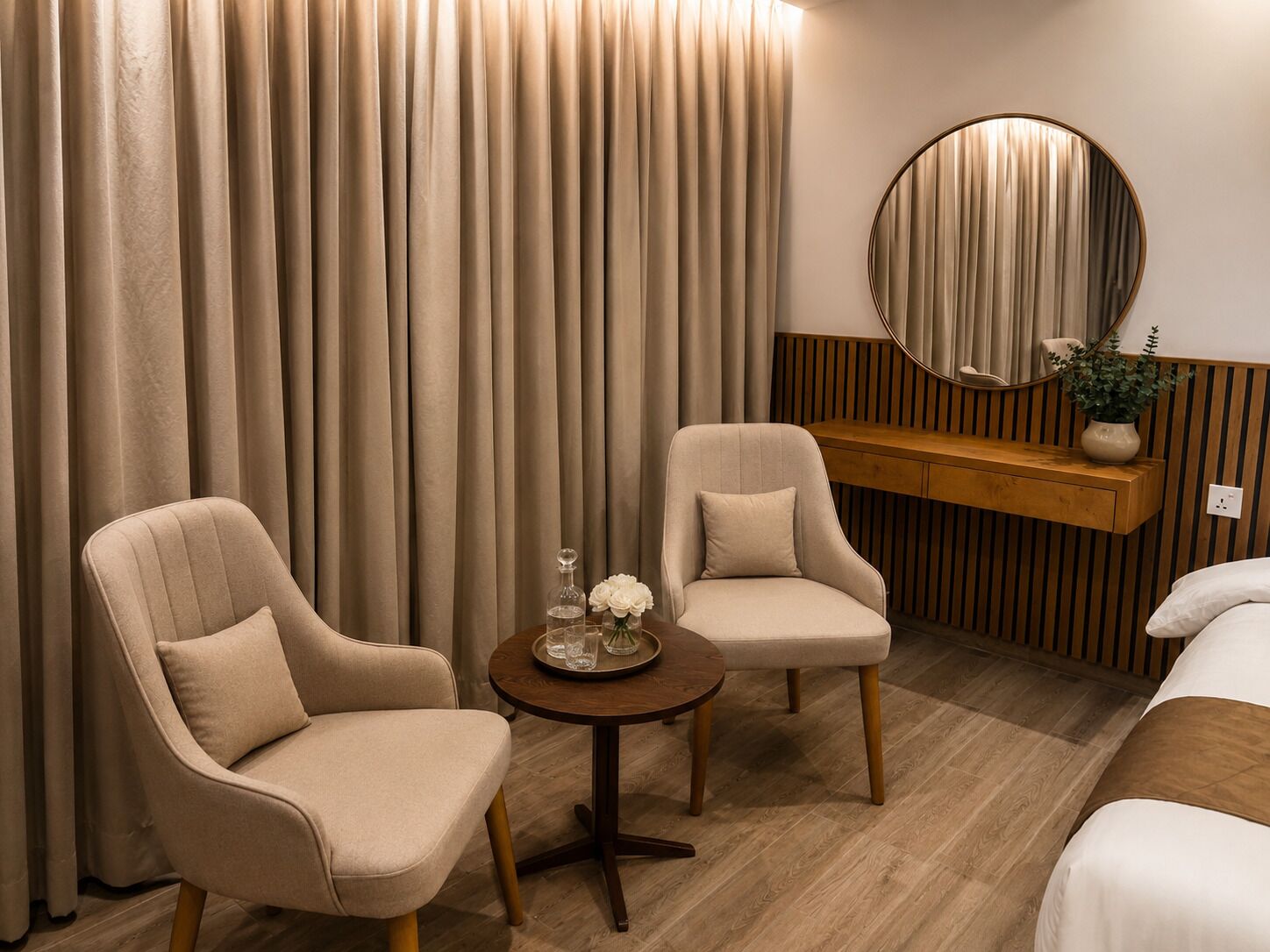 A hotel room with two armchairs, a small round table with a vase and glassware, a wooden console table, and a round mirror on the wall.