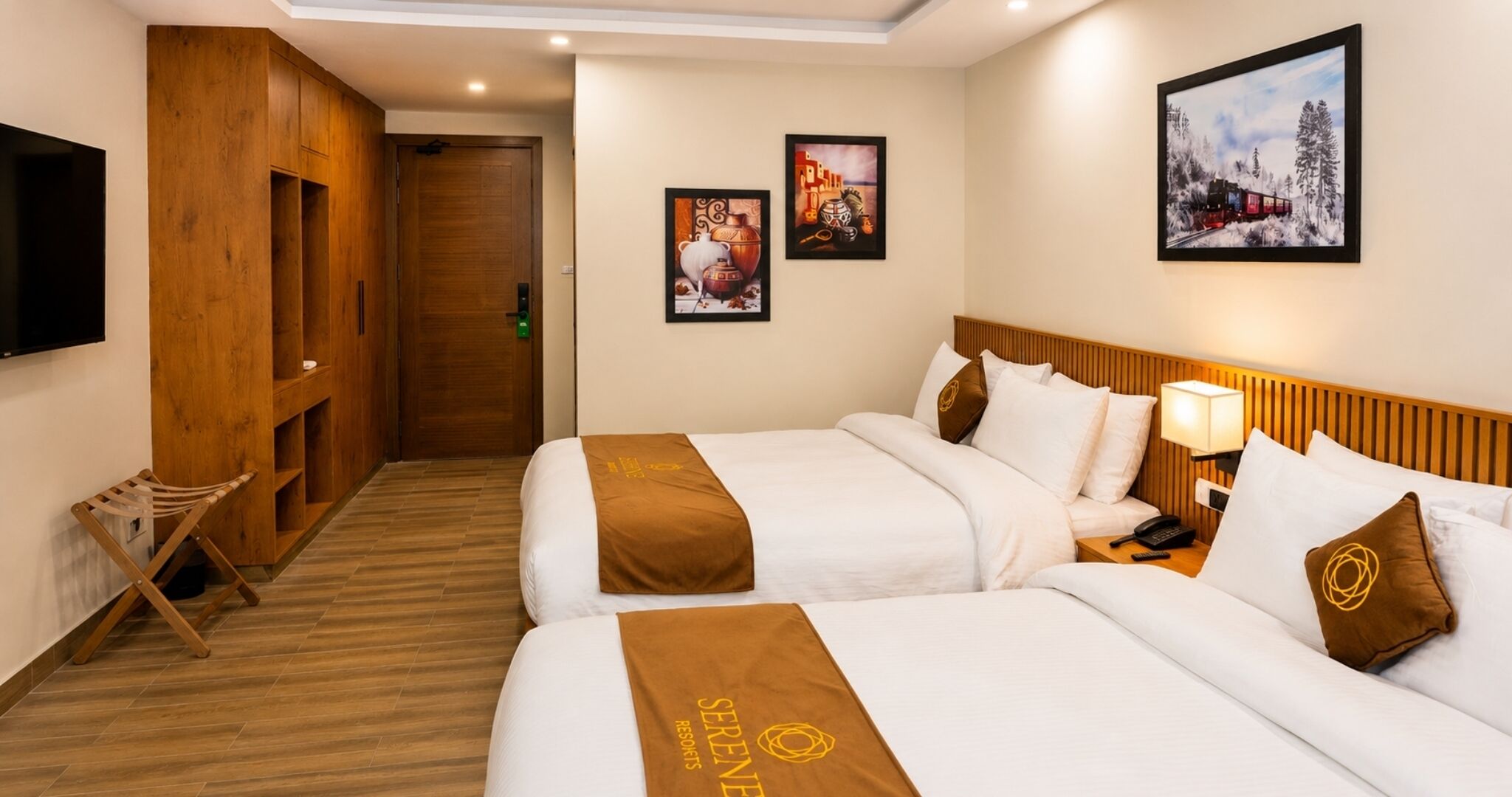 A hotel room with two beds, wooden headboards, a TV, artwork on the walls, and a small seating area.