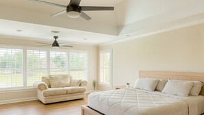 Ein Schlafzimmer mit einem großen Bett, einem Sessel, einem Deckenventilator und großen Fenstern.