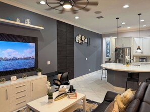 A modern living room with a TV, seating, a kitchen area, and a dining space. - Fully Themed 5BR w/ Pool + Arcade | Resort Access! (Davenport)