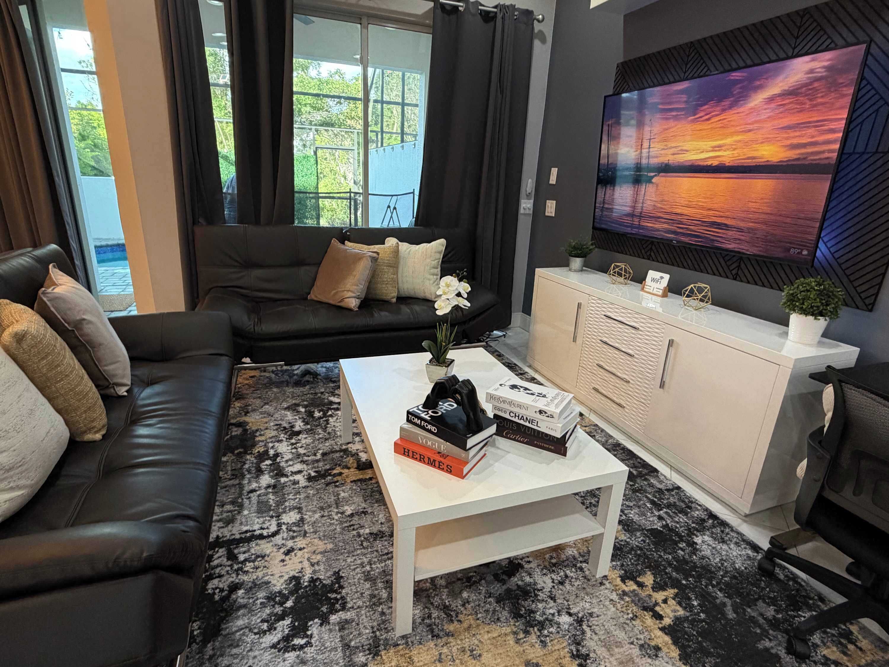 A modern living room with a black leather sofa, a white coffee table with books, a white cabinet, and a large TV displaying a sunset view.