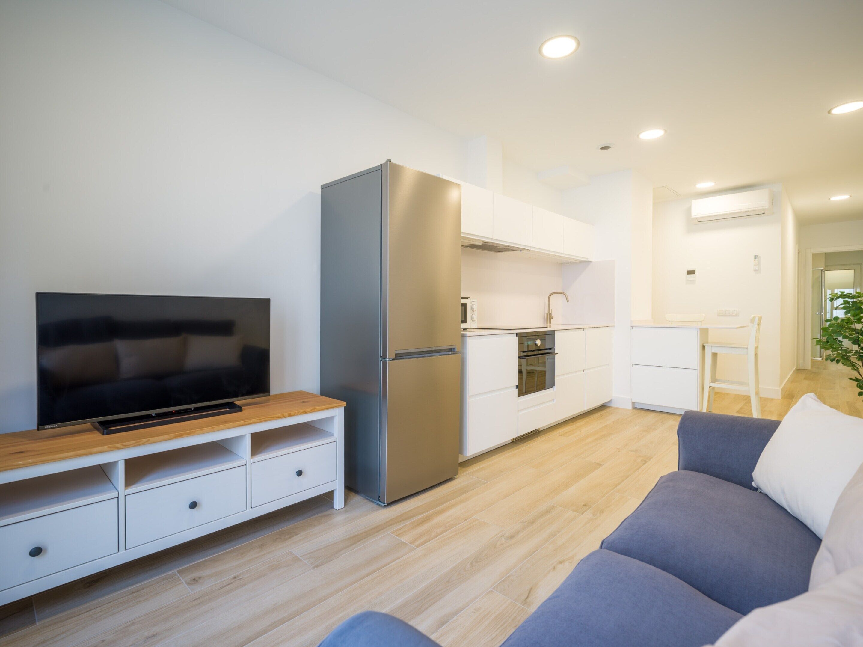 A modern living room with a flat-screen TV, a sofa, and a kitchen area with a refrigerator and oven.