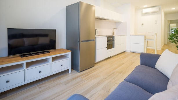 A modern living room with a flat-screen TV, a sofa, and a kitchen area with a refrigerator and oven. - Deluxe Canteras 2C By Canariasgetaway (Las Palmas de Gran Canaria)