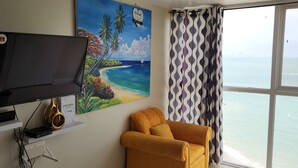 A room with a large window, a painting of a beach, a yellow sofa, and a television on a shelf. - Scenic studio in Portmore, Jamaica (Kingston)